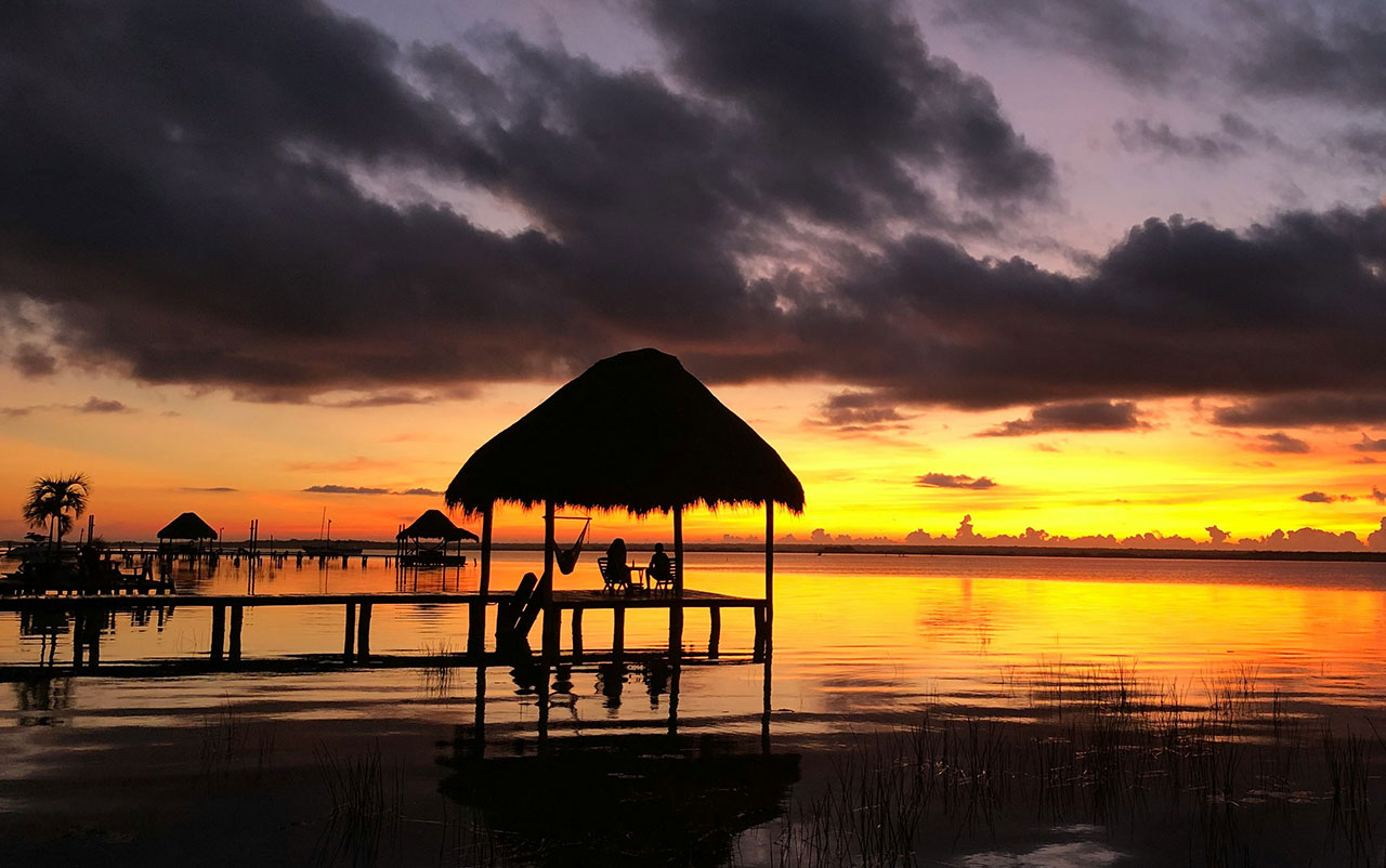A thatched-roof palapa pier sits on the calm water of a lagoon during a vibrant orange and purple sunset.