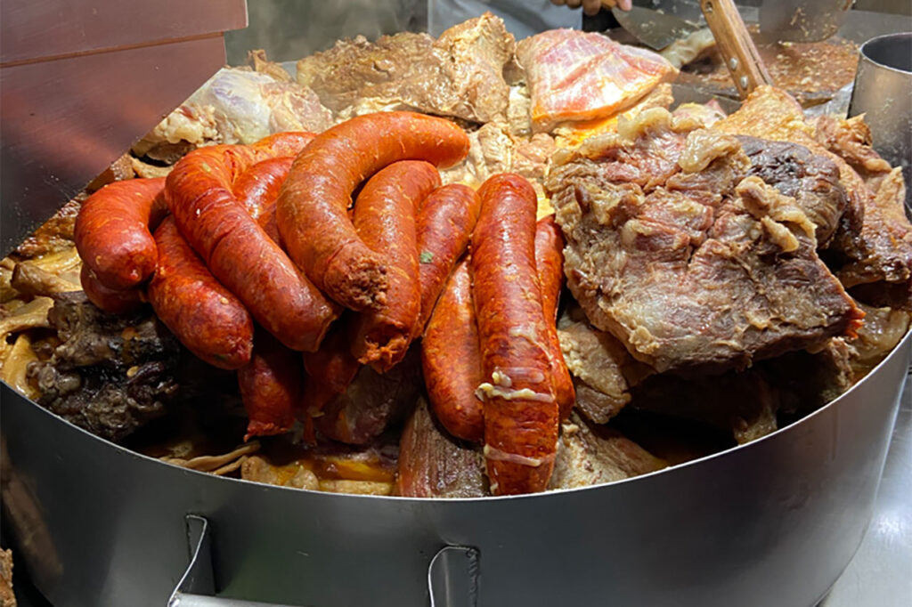 A large metal pan filled with a variety of meats including suadero, longaniza sausages, and tripe cooking in their own fat at a traditional Mexico City street food stand.
