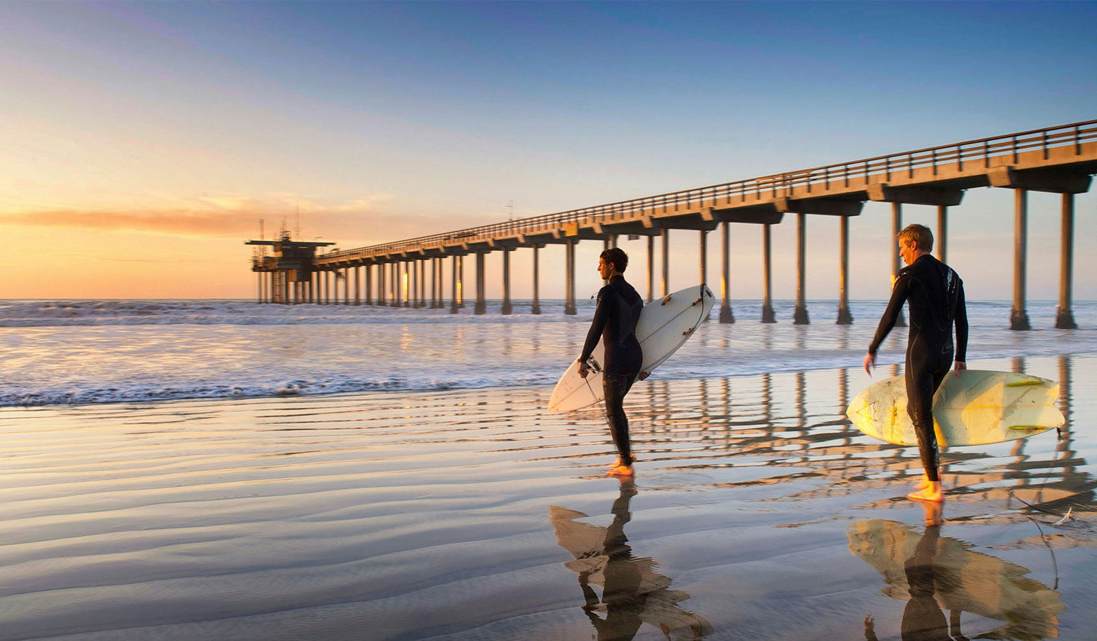 Two surfers carrying boards walk along the wet sand toward a long wooden pier during a hazy morning at Mission Beach.