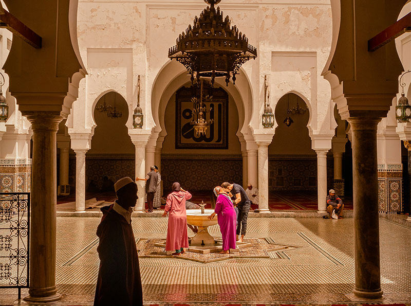 Worshippers at a star-shaped fountain inside an ornate Moroccan courtyard featuring white pillars, archways, and a large chandelier.