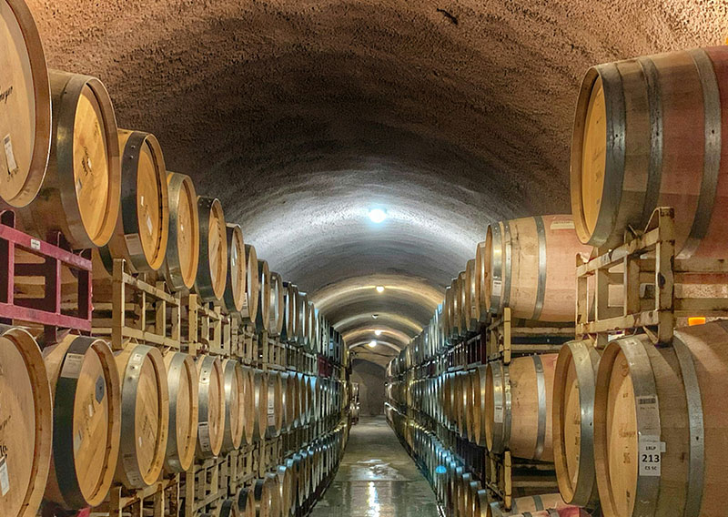 A long, symmetrical view down a dimly lit underground wine cave in Napa Valley, with rows of oak wine barrels stacked on wooden racks lining both sides of a stone-arched corridor.