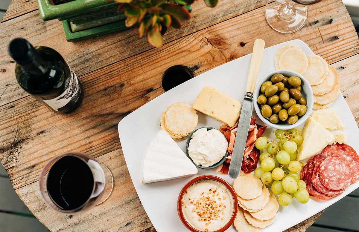 A close-up overhead shot of a white plate featuring a wine country picnic spread, including wedges of artisan cheese, sliced deli meats, a small bowl of green olives, a cluster of green grapes, and a small dish of red pepper jelly or jam