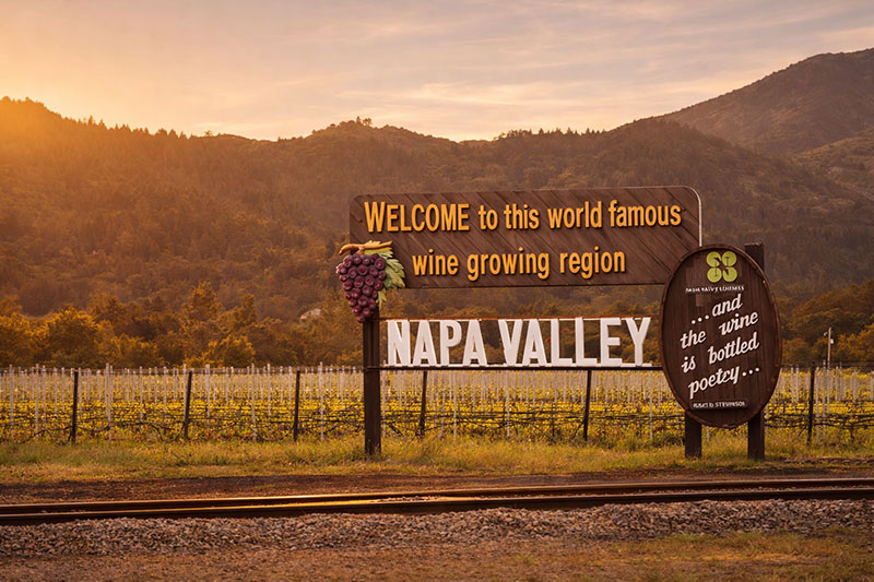 The iconic "Welcome to this world famous wine growing region Napa Valley" wooden sign standing at the edge of a vineyard, captured at sunset with the hills in the background under a warm, golden sky.