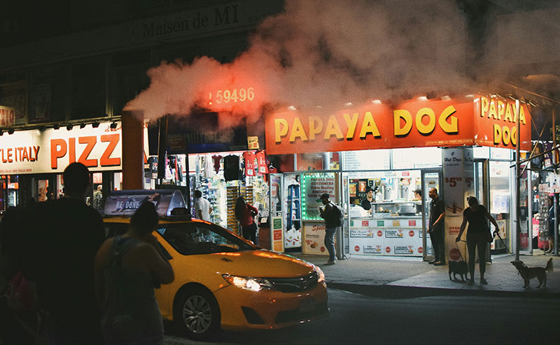 A yellow New York City taxi cab drives past a "Papaya Dog" storefront at night, with glowing neon signs advertising hot dogs and pizza slices in a busy Manhattan neighborhood.