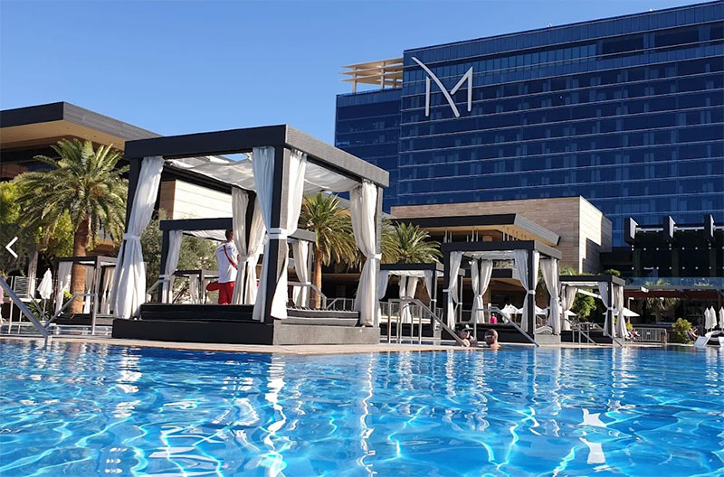 An exterior view of the M Resort Spa Casino in Henderson, featuring the modern blue glass hotel tower and the large outdoor pool deck with lounge chairs and cabanas.