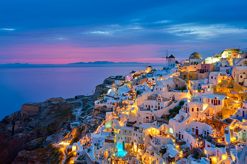A panoramic sunset view of Oia, Santorini, featuring traditional white-washed buildings and blue-domed churches nestled on the volcanic cliffs overlooking the Aegean Sea.