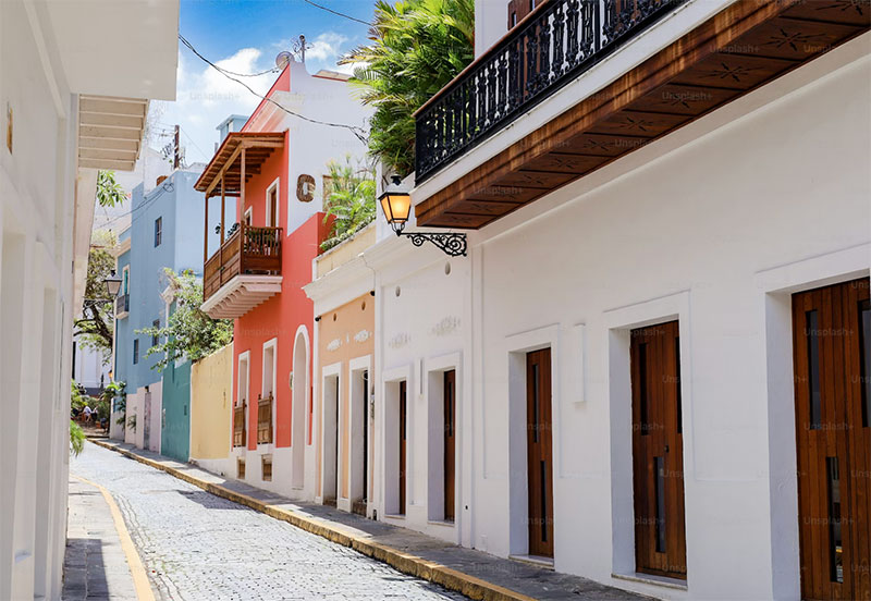 A bright, sunlit cobblestone street in Old San Juan lined with colorful colonial buildings and balconies.