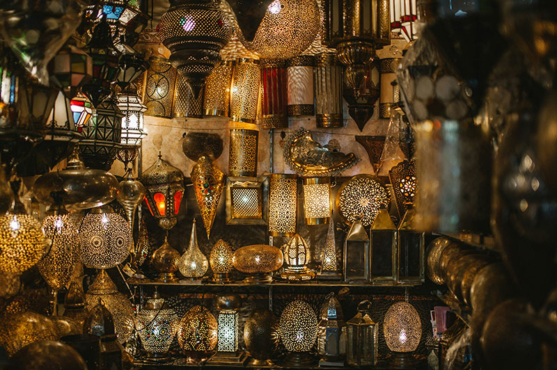 A dark shop stall filled with glowing, intricately pierced metal Moroccan lanterns of various shapes and sizes.