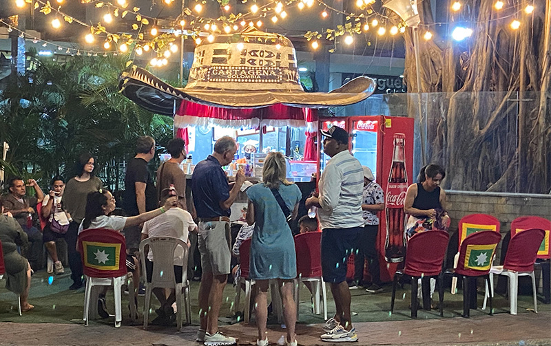 Locals and tourists gathering for fresh oysters and ceviche at Ostreria Del Mar Rojo, an outdoor food stand shaped like a giant Sombrero Vueltiao under string lights in Cartagena.