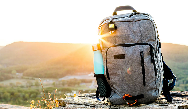 A structured, gray carry-on travel backpack standing upright outdoors in warm, golden-hour sunlight.