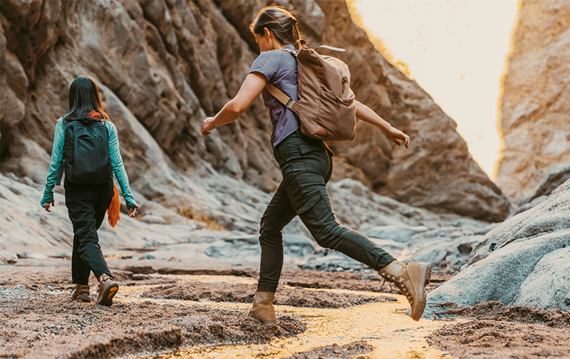 A woman wearing a brown backpack and olive green pants walks confidently across a rocky, sunlit desert landscape. She is captured mid-stride, highlighting a sense of movement and adventure against the rugged terrain.