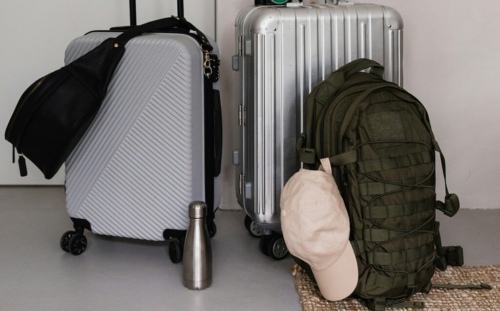 An overhead shot of two pieces of luggage resting against a wall: a silver hard-shell roller suitcase and a dark olive green travel backpack with a tan baseball cap hanging from the strap. Both bags are compact, illustrating a streamlined two-bag travel setup.