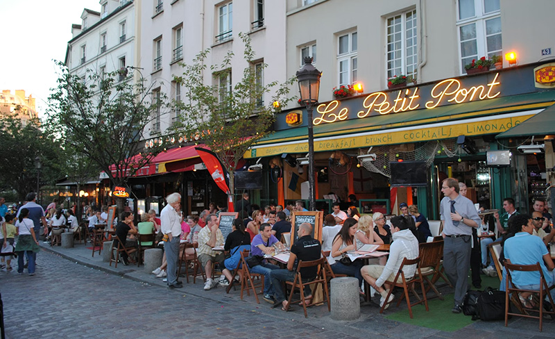 The red awning and outdoor seating of Le Petit Pont cafe in the Latin Quarter, with patrons dining at small round tables on a narrow Parisian street.