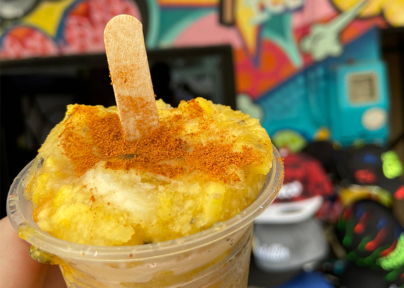A close-up of a passion fruit popsicle (crema de maracuyá) in a clear plastic cup, featuring a creamy yellow texture topped with fresh passion fruit pulp, seeds, and a dusting of cinnamon, with a wooden stick inserted.