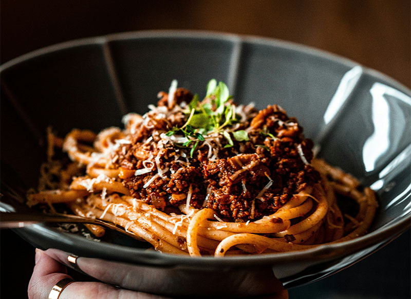 A close-up shot of a person holding a dark gray ceramic bowl filled with thick pasta topped with a rich meat ragù. The dish is garnished with freshly grated cheese and a sprig of fresh green herbs. The lighting highlights the glossy texture of the sauce and the steam coming off the warm meal.
