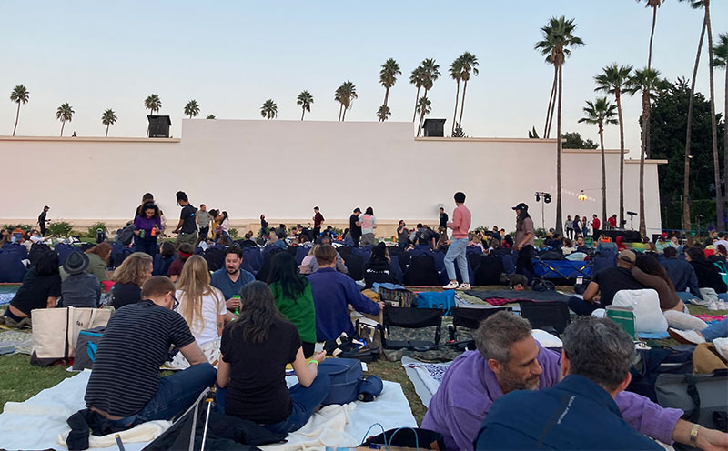 A large crowd of people sitting on blankets with picnic setups on the Fairbanks Lawn at Hollywood Forever Cemetery during dusk.