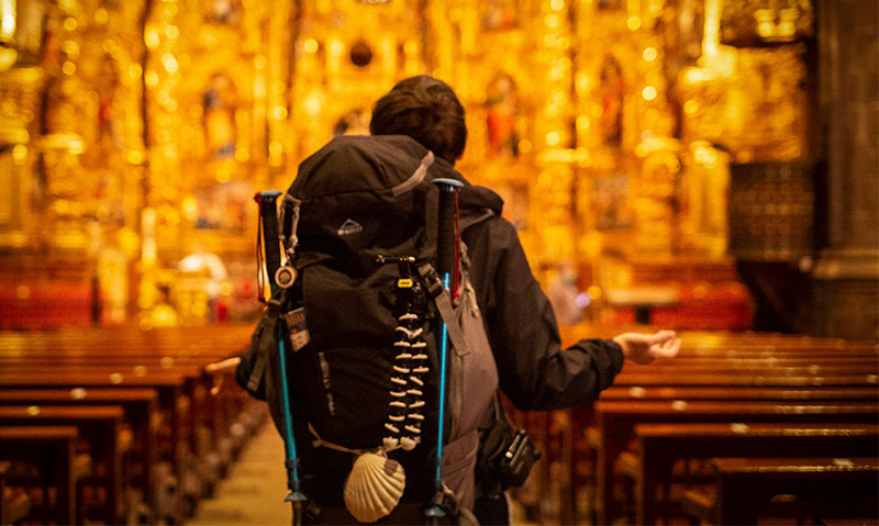 A pilgrim wearing a backpack and carrying trekking poles stands inside a dimly lit historic Spanish church with a golden altarpiece.