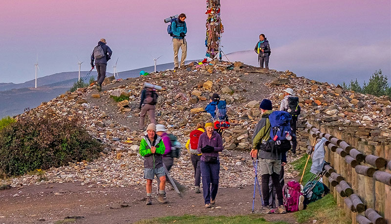 Pilgrims with backpacks resting at the Cruz de Ferro, a large pile of stones and a wooden cross at sunrise on the Camino Francés.