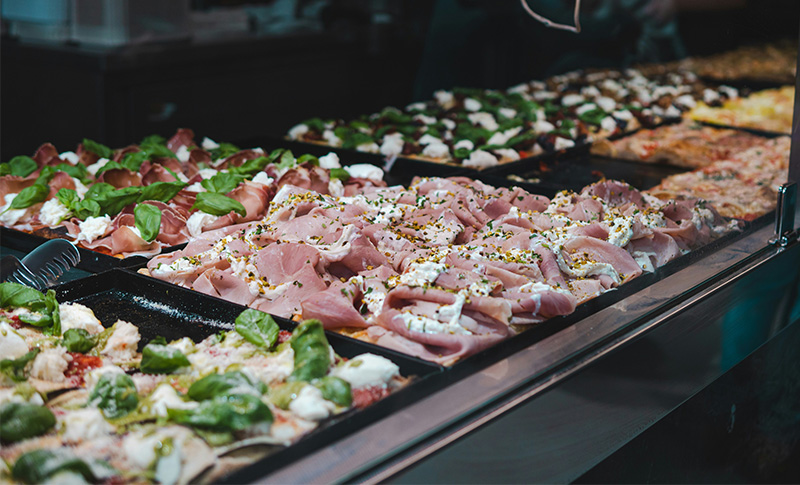 A bright, eye-level shot of a modern Roman pizza shop window displaying several long, rectangular trays of pizza al taglio. The varieties include a classic Margherita with vibrant red tomato sauce and melted white mozzarella, a white pizza topped with thinly sliced potatoes and rosemary, and another with bright green zucchini flower