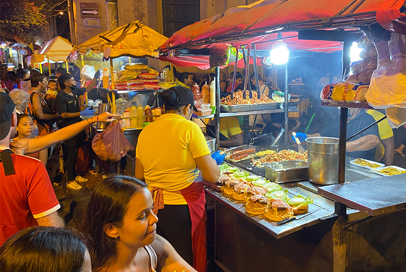 A busy street food vendor preparing hamburgers on a flat top grill at a brightly lit night cart in Plaza de la Trinidad