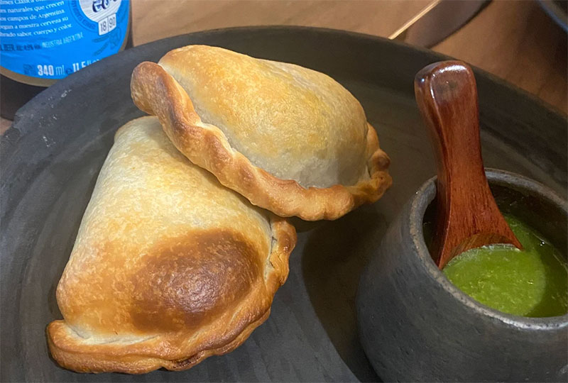 Two golden-brown baked empanadas filled with pork and beef served next to a dark cup of spicy green llajua sauce.
