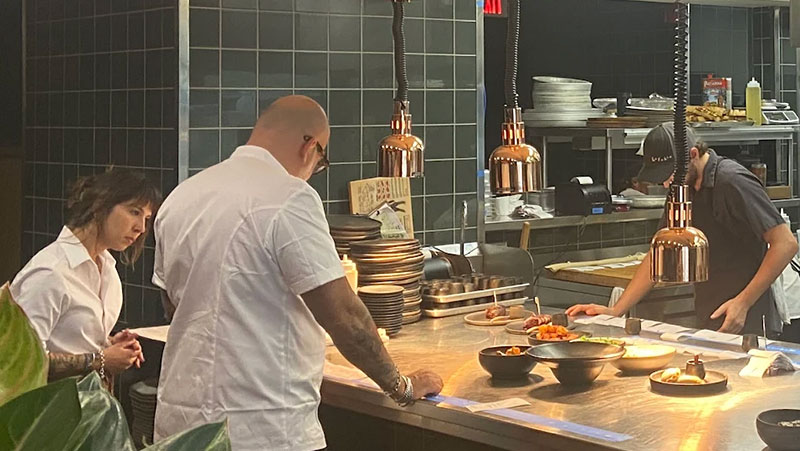 Chef Daniel del Prado standing in the open kitchen at Porzana expediting plates under copper warming lamps.