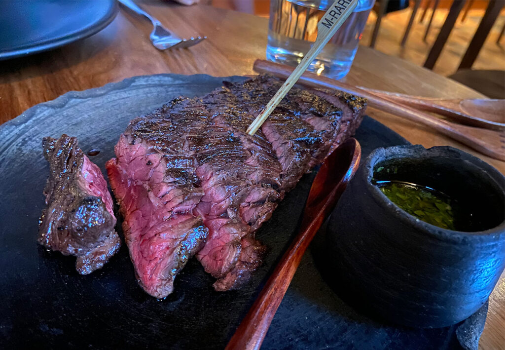A perfectly medium-rare sliced Entraña skirt steak served on a dark stone plate with a side of green chimichurri sauce.