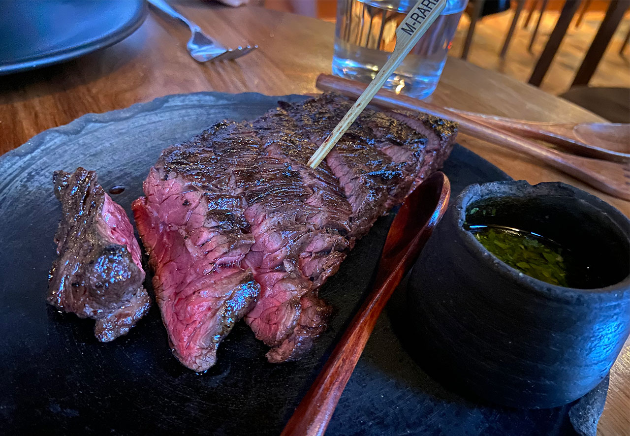 A perfectly medium-rare sliced Entraña skirt steak served on a dark stone plate with a side of green chimichurri sauce.
