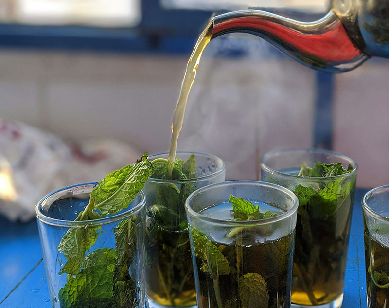 Steaming hot tea being poured from a silver teapot into small glass cups generously filled with fresh green mint leaves.