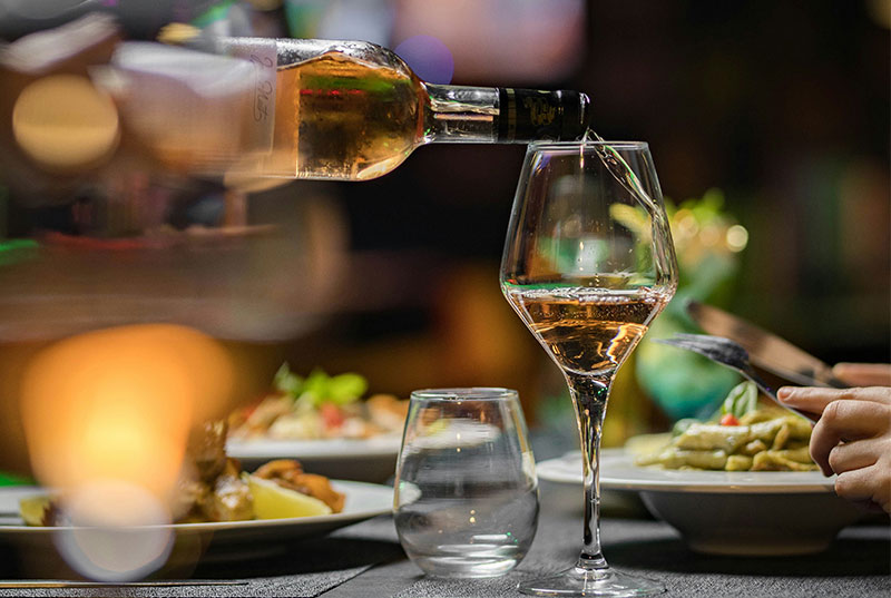 A close-up of rosé wine being poured into a glass at a dinner table with blurred plates of food in the background.