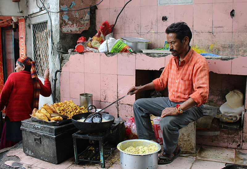 A street food vendor in an orange shirt sits by a large wok of hot oil, frying traditional snacks at a pink-tiled stall in India.