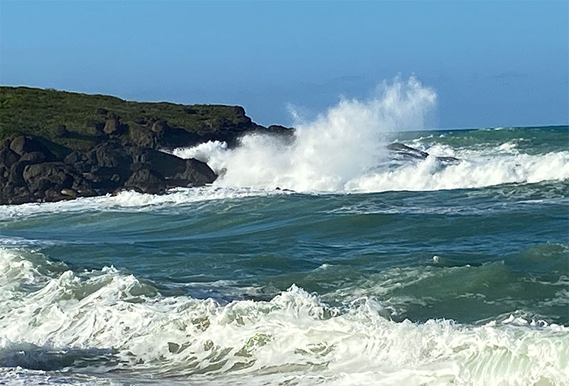 Powerful turquoise waves crashing onto a sandy beach with rocky green cliffs in the distance.
