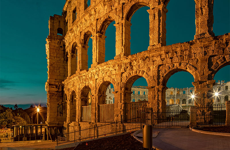 The towering stone arches of the ancient Roman Pula Arena illuminated at night against a dark sky in Istria, Croatia.