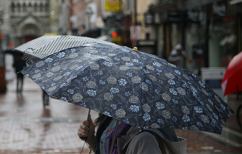 A rainy day scene on a busy Dublin street, featuring pedestrians with colorful rain jackets walking past historic brick buildings and a traditional blue storefront, with the wet pavement reflecting the city lights.