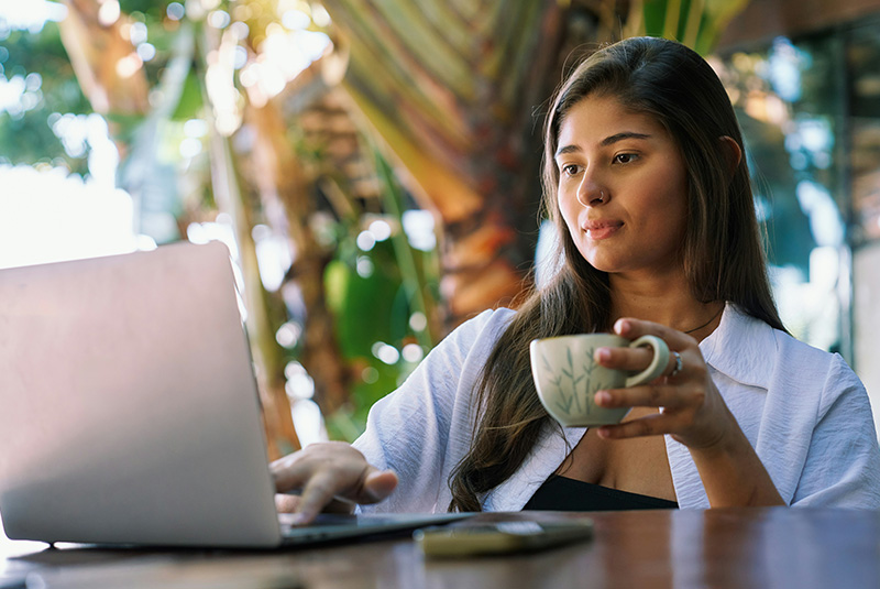 A young woman works on her laptop while holding a coffee mug at a lush, open-air cafe.