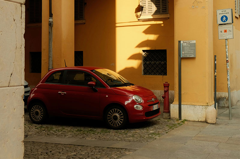 A small red rental car parked on a cobblestone street in the historic center of Modena, Italy, with traditional Italian architecture in the background.