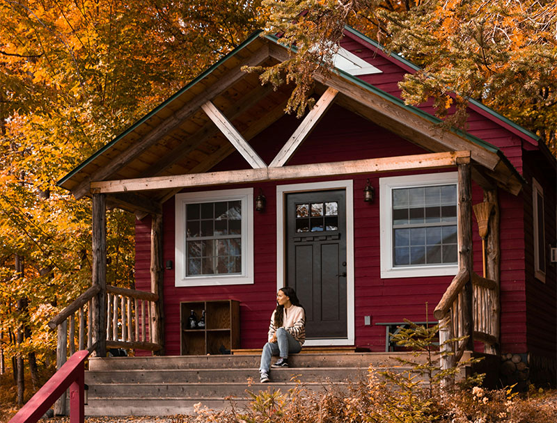 A cozy, A-frame wooden cabin tucked into a dense forest of tall evergreen trees, with a warm light glowing from the windows and a light dusting of snow on the surrounding ground.