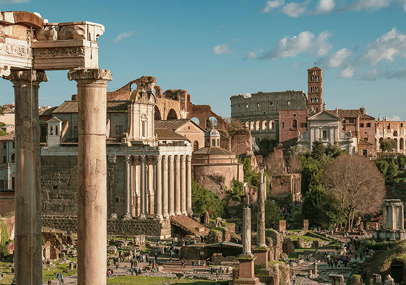 A wide-angle, slightly elevated shot of the Roman Forum at sunrise. In the foreground, the weathered stone columns of the Temple of Saturn stand tall against a soft pink and orange sky.