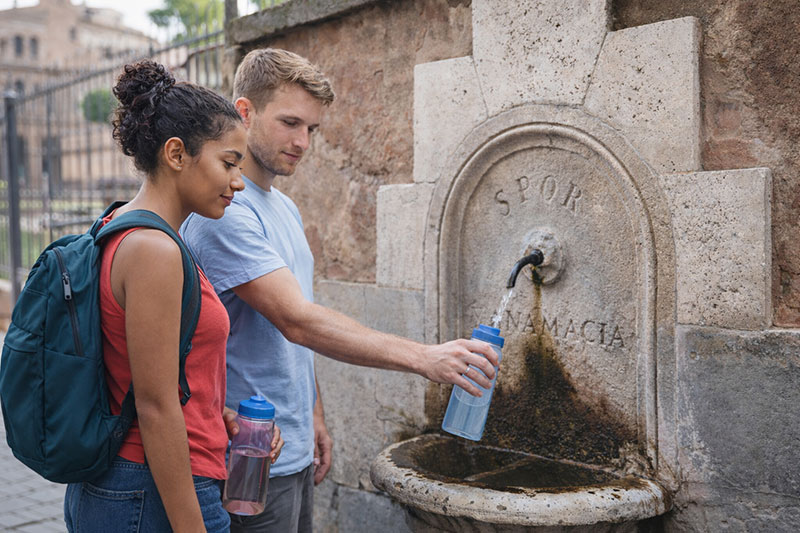 Young couple travelers refilling reusable water bottles at a public fountain in Rome while sightseeing