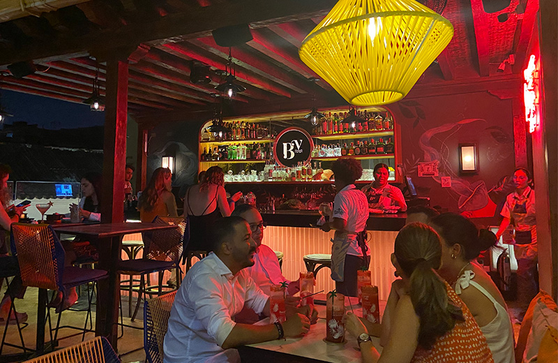 A vibrant rooftop bar at night with people sitting at tables under a large yellow woven lamp and red ambient lighting at Buena Vida Marisqueria