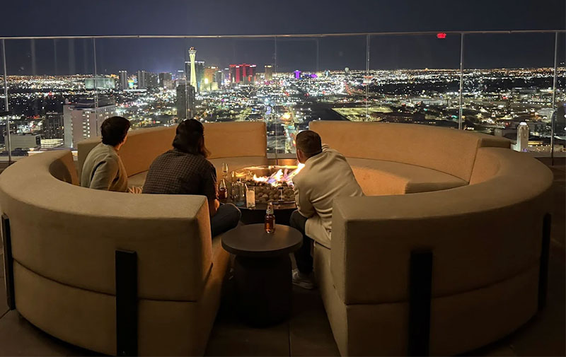 A nighttime view of two cocktails on a table at the Legacy Club rooftop bar, with the glowing lights of the Las Vegas skyline and the STRAT Hotel visible in the background.