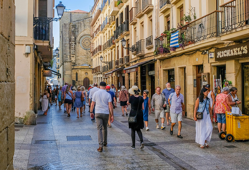 Alt Text A narrow, sunlit pedestrian walking street in the Old Town of San Sebastián, Spain, lined with multi-story historic buildings featuring wrought-iron balconies and ground-floor shops, with people strolling in the distance.