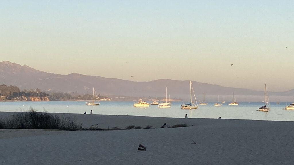 A wide scenic view of the Santa Barbara harbor at golden hour, with several sailboats anchored in the calm blue water and a paddleboarder in the foreground.