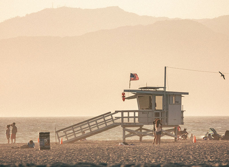 A classic wooden lifeguard tower with an American flag on the sand at Santa Monica beach with hazy mountains in the background.