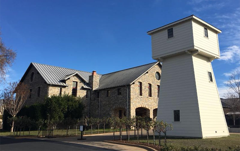 The exterior of Silver Oak Cellars in Napa Valley, featuring the iconic stone water tower and rustic stone winery buildings set against a backdrop of green vineyards and a bright blue sky.