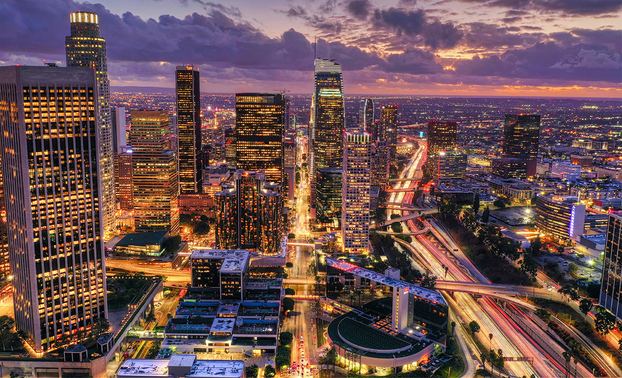 A wide night skyline view of Los Angeles featuring illuminated skyscrapers and glowing city lights under a dark sky.