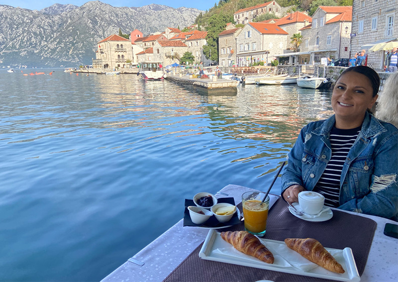 A woman smiles at a waterfront restaurant table in Montenegro, with a breakfast of coffee and croissants set against a backdrop of historic stone buildings and mountains.