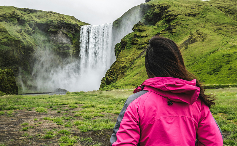 A solo female traveler wearing a bright pink jacket stands in a green field, looking up at a towering, powerful waterfall.