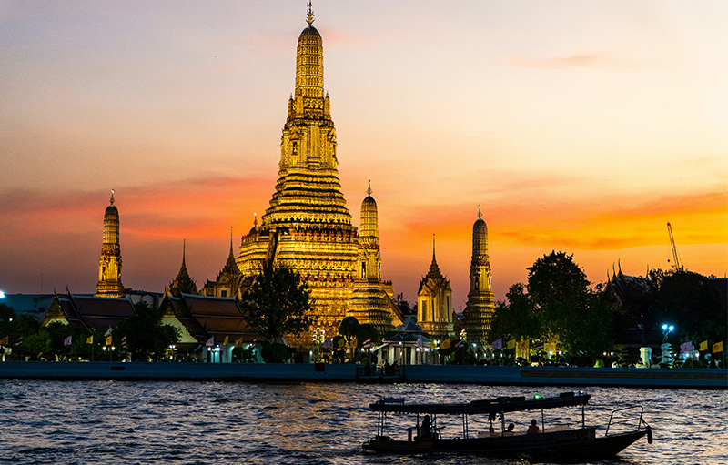 A golden Buddhist temple with intricate spires (Wat Arun) reflecting in a river during a vibrant orange and yellow sunset in Bangkok, Thailand.