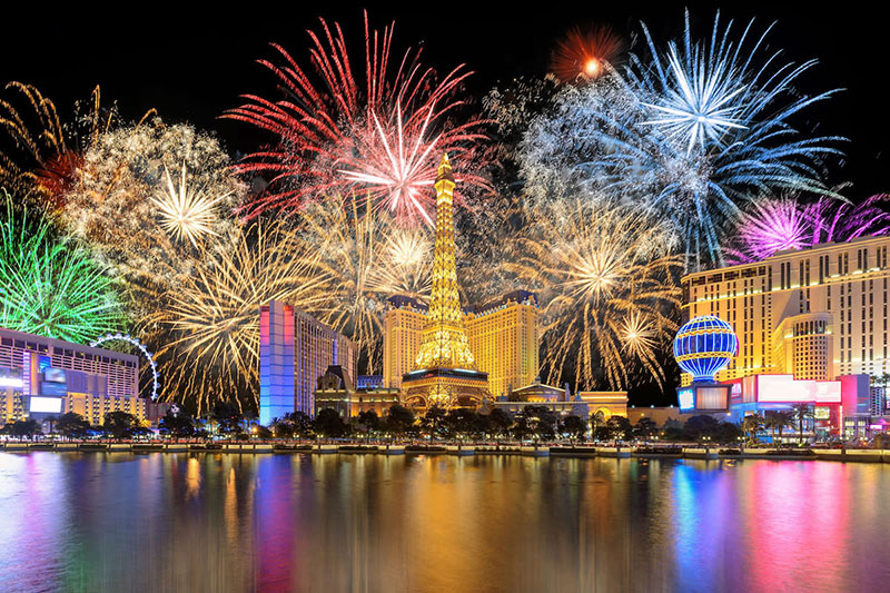 A wide-angle night shot of spectacular New Year’s Eve fireworks exploding in bursts of white and gold over the Las Vegas Strip, reflecting in the water in the foreground.
